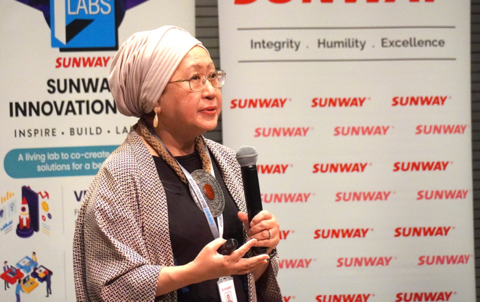 Prof. Tan Sri Dr. Jemilah holding a microphone and a clicker, giving a presentation amidst the backdrop of Sunway University and DHL bunting