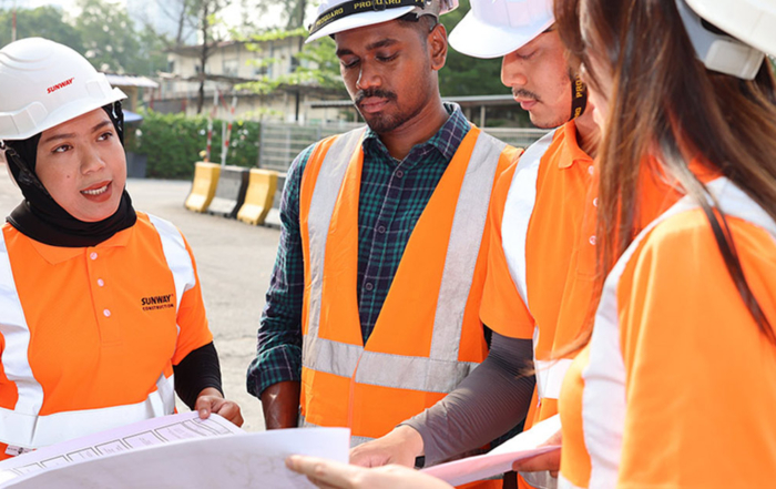 Four Sunway Construction team members of various races, clad in Sunway Construction orange reflector vest and Sunway hard hats at a construction site 